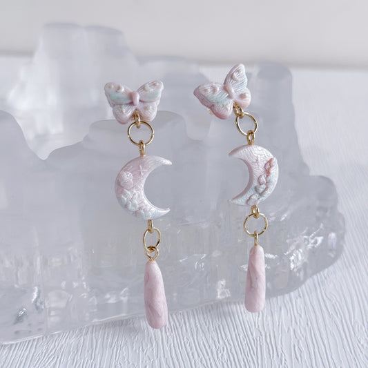 Pair of earrings with butterfly and moon designs on a clear stand.